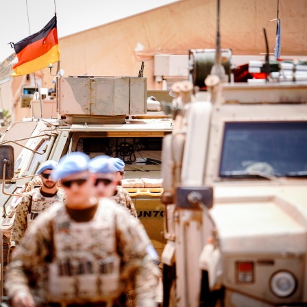 Die Deutschlandfahne und die Flagge der Vereinten Nationen wehen im Camp Castor in Gao, im Vordergrund sind Soldaten und gepanzerte Fahrzeuge zu sehen.