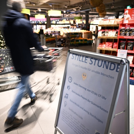 Ein Mann schiebt seinen Einkaufswagen in den Supermarkt, während im Vordergrund ein Schild auf die Stille Stunde im Edeka hinweist.