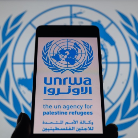 Das Logo des UNRWA, des Hilfswerd der Vereinten Nationen für Palästina-Flüchtlinge, ist auf einem Smarphone-Bildschirm zu sehen, im Hintergrund erkennt man das Logo der Vereinten Nationen.