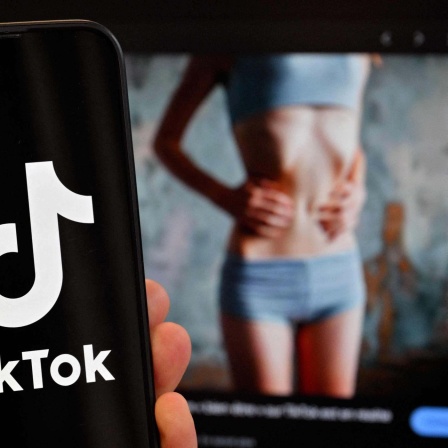 Logo der chinesischen Social-Media-Anwendung TikTok auf einem Smartphone vor einem Bildschirm, auf dem das Bild einer dünnen Frau angezeigt wird. 