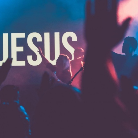 Ein Pop-Konzert in einer Kirche, mit blauem Licht und tanzenden Silhouetten. Im Hintergrund strahlt ein weißer "Jesus"-Schriftzug.