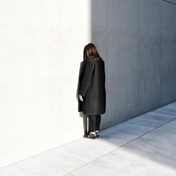 Eine Person mit schwarzer Kleidung steht mit gesenktem Kopf direkt vor einer grauen Wand.