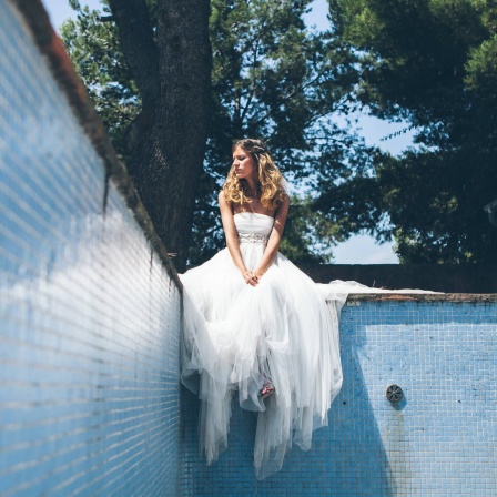 Eine junge Frau im weißen Brautkleid sitzt am Rand eines leeren Schwimmbeckens und sieht mäßig glücklich aus
