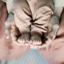 Die Füße eines Babys werden von den Händen einer erwachsenen Person auf dem Schoß gehalten.