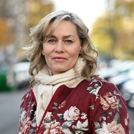 Die Schauspielerin Gesine Cukrowski steht bei einem Fototermin in ihrem Kiez. Sie trägt eine geblümte Jacke und einen beigen Schal. Der Hintergrund ist verschwommen.