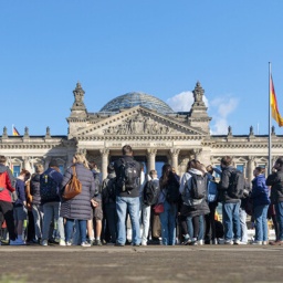 Symbolbild Deutscher Bundestag Deutschland, Berlin , Platz der Republik, der Reichstagbau in der Herbstsonne mit blauem Himmel und weißen Wolken, wehender Deutschlandfahne. Im Vordergrund junge Menschen in Rückenansicht, die sich interessiert dem Reichstag zuwenden.