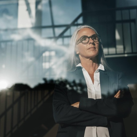 Eine Frau im schwarzen Blazer steht mit verschränkten Armen an einem Fenster und schaut mit entschlossenem Blick hinaus.