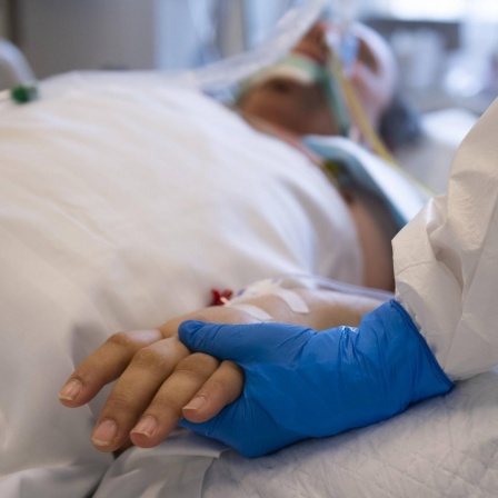 Ein medizinischer Mitarbeiter hält die Hand eines Patienten der im Krankenbett liegt mit einem Beatmungsgerät.