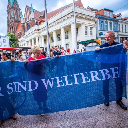 Manuela Schwesig (M, SPD), Ministerpräsidentin von Mecklenburg-Vorpommern, Rico Badenschier (l, SPD), Oberbürgermeister von Schwerin, und der Vorsitzende des Welterbefördervereins, Joachim Brennecke, halten nach der Entscheidung zur Aufnahme des Schweriner Residenzensembles in die Welterbeliste eine Fahne mit der Aufschrift "Wir sind Welterbe".