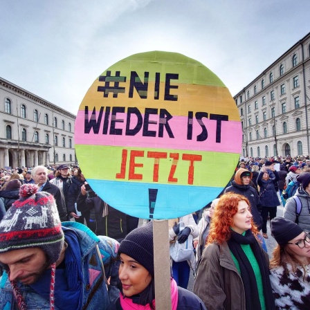 Eine Menschenmenge in einer Münchener Straße, im Vordergrund ein buntes rundes Plakat mit der Aufschrift "#Nie wieder ist jetzt"