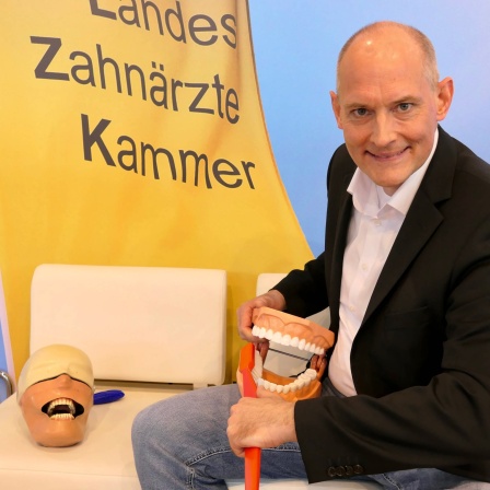 Zahnarzt Elmar Ludwig sitzt vor einem Banner mit der Aufschrift &#034;LandesÄrzteKammer&#034; und putzt mit einer großen Zahnbürste einem übergroßen Gebiß die Zähne. In SWR1 Leute erklärt er, wie gute Mundhygiene bis ins hohe Alter aussieht.