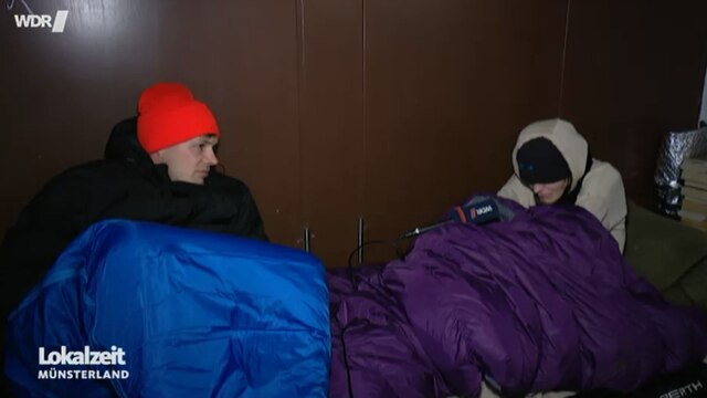 Zwei Männer sitzen in einem Schlafsack auf der Straße mit einem Mikrofon.