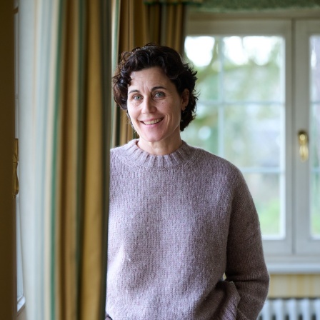 Schauspielerin Nina Kunzendorf steht mit einem rosa Pulli und blauer Jeans an einem Fenster