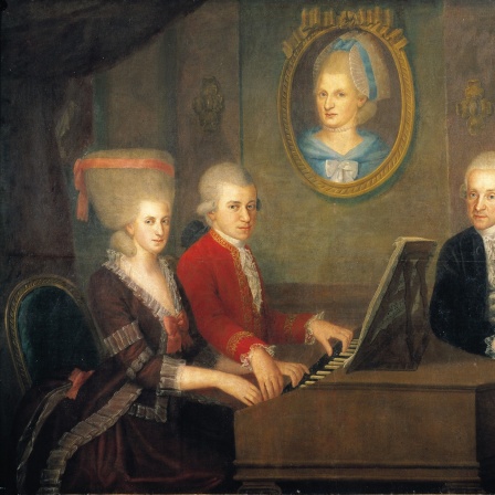 An einem Flügel sitzen Nannerl und Wolfgang Mozart, der Vater steht mit Geige hinter dem Instrument. Und die Mutter ist als Gemälde an der Wand in die Szene eingearbeitet.