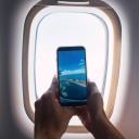 Ein Mann, von dem man nur die Hände sieht, macht mit dem Smartphone ein Foto durch das Fenster eines Flugzeugs.