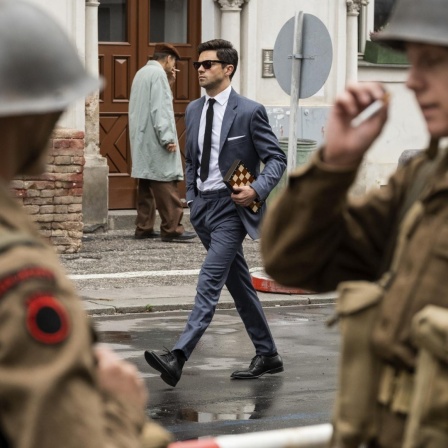 Szenenfoto aus der Spionage-Serie "Spy City", auf dem der Schauspieler Dominic Cooper in der Rolle des Agenten Fielding Scott mit einem gefalteten Schachbrett unter dem Arm an zwei Soldaten vorbei die Straße hinunter läuft.