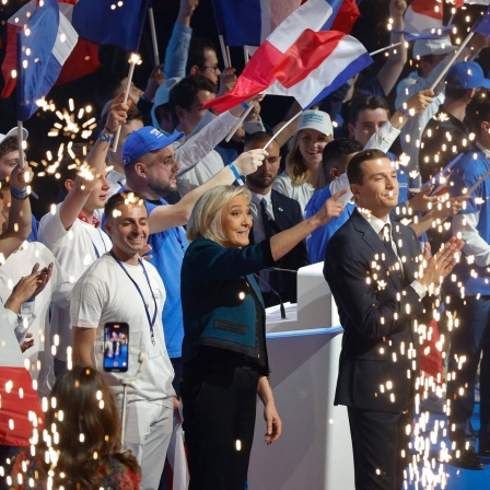 Marine Le Pen und ihr Parteichef Bardella vom Rassemblement National stehen auf einer Bühne, umringt von Anhängern, und feiern.
