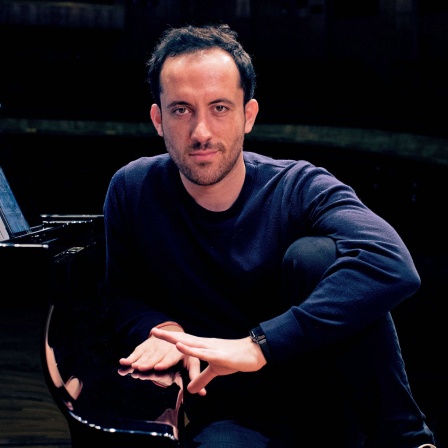 Igor Levit, Mann in dunkler Kleidung vor dunklem Hintergrund am Klavier