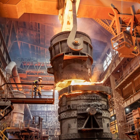 Ein großes, futuristisch aussehendes Stahlwerk, in dessen Mitte ganz klein ein Mann auf einer Brücke steht und zusieht wie flüssiger, heißer Stahl in einen großen Kessel fließt