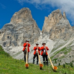 Drei Alphornbläser in Trachtenanzügen stehen vor aufregendem Gebirge auf einer Wiese.