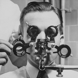 Eine historische schwarz-weiß Fotografie zeigt den Kopf eines Mannes hinter einem Augenmessgerät in einen Klinik.