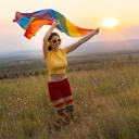 Glückliche junge Frau in sommerlicher bunter Kleidung mit Kopfhörern und Regenbogenfahne vor einem Sonnenuntergang.