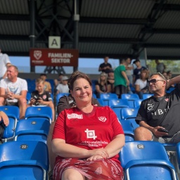 Frau mit roten Kleid sitzt in einem Fußballstadion mit blauen Sitzen, unter weiteren Zuschauern.