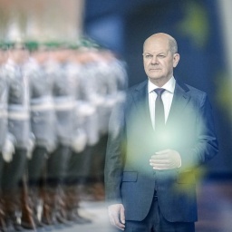 Bundeskanzler Olaf Scholz (SPD) wartet hinter einer Spiegelung mit der Europa-Fahne und einem Bundeswehr-Corps vorm Bundeskanzleramt auf einen Staatsgast.
