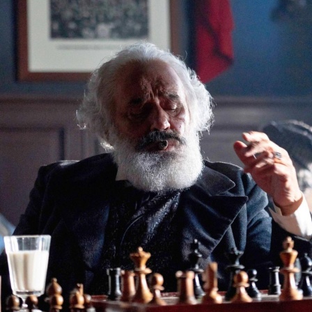 Mario Adorf, in der Rolle des Karl Marx, sitzt mit einem Schachbertt und einem Glas Alkohol am Tisch.