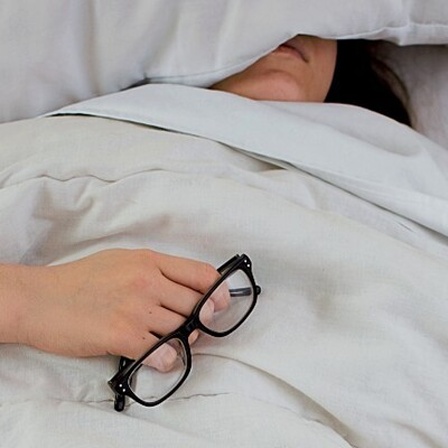 Eine Frau liegt im Bett, den Kopf unterm Kissen und die Brille in der Hand.