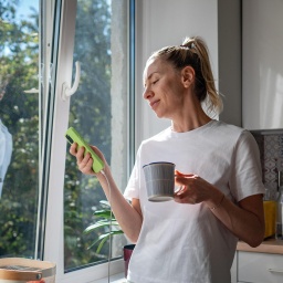 Eine Frau steht am Morgen entspannt in der Küche mit Kaffeetasse und Handy