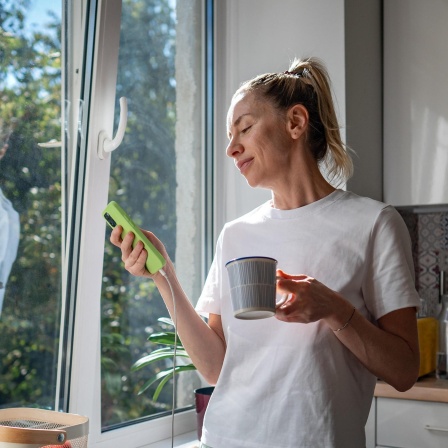 Eine Frau steht am Morgen entspannt in der Küche mit Kaffeetasse und Handy