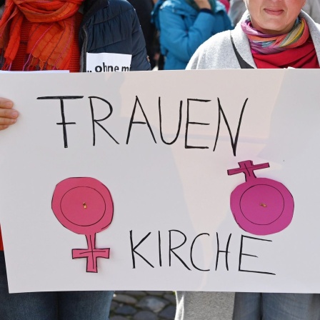 Frauen halten ein Plakat mit der Aufschrift "Frauen Kirche" und dem Symbol für Weiblichkeit, bei dem das Kreuz zusätzlich markiert ist.