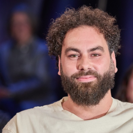 Autor Dominik Bloh, der früher obdachlos war, sitzt in einem Fernsehstudio und blickt freudlich in die Kamera. Er hat braunes krauseliges Haar und einen passenden Bart dazu. Er trägt ein beiges T-Shirt.