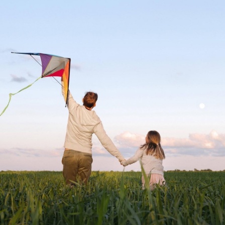 Ein Mann hält einen Drachen, während er mit seiner Tochter an der Hand durch ein Feld läuft.