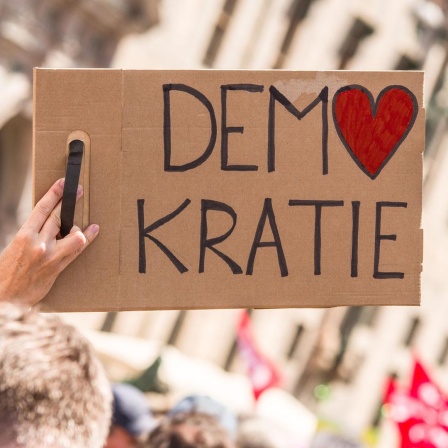 Bei einer Kundgebung wird ein Schild mit der Aufschrift "Demokratie" in die Höhe gestreckt. Dabei ist das O in Demokratie in Form eines Herzes.