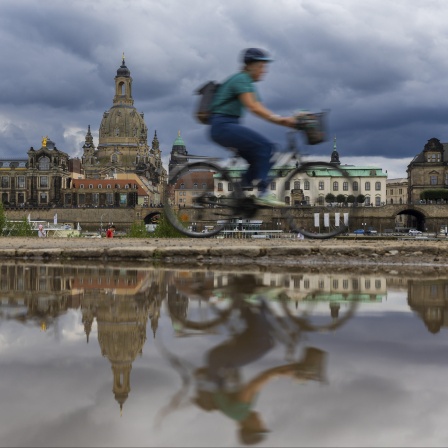 Die Silhouette eines Radfahrers sowie die im Hintergrund befindlichen dunklen Wolken und die Frauenkirche spiegeln sich in einer Pfütze.