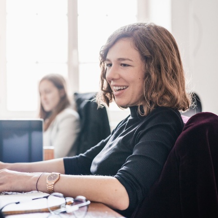 Eine Frau sitzt in einem Büro vor einem Computerbildschirm und lacht. 