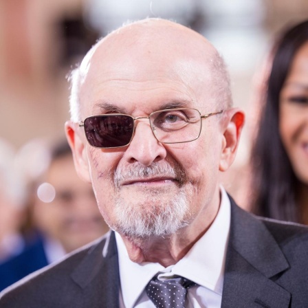 Porträt von Salman Rushdie mit dunklem Anzug und Krawatte in weissem Hemd. Im Hintergrund sind Personen verschwommen zu sehen. Das Brillenglas vor dem rechten Auge ist geschwärzt.