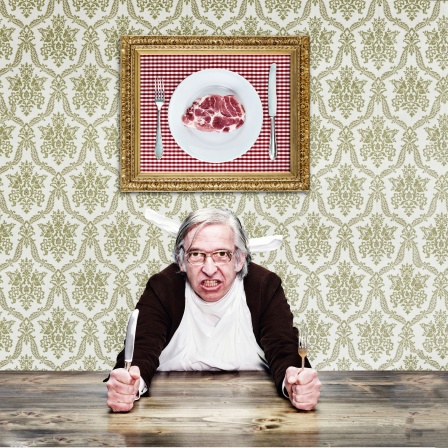 Ein Mann mit grauem Haar und Brille am Tisch, hält Messer und Gabel in den Händen, dahinter ein Foto mit Fleisch an der Wand.