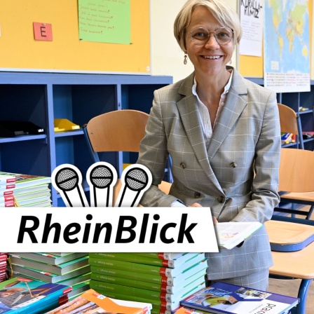 NRW-Schulministerin Dorothe Feller (CDU) sitzt in einem leeren Kalssenraum und hat hinter einen Stapel Schulbücher ein Buch aufgeschlagen