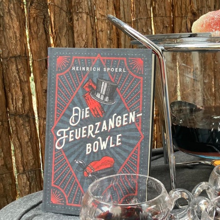Das Buch von Heinrich Spoerl "Die Feuerzangenbowle" steht auf einem Tisch neben einer Feuerzangenbowle im Freien