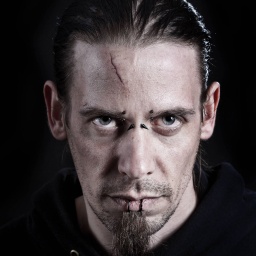 Dunkel gekleideter Mann mit Piercings und Narbe auf der Stirn