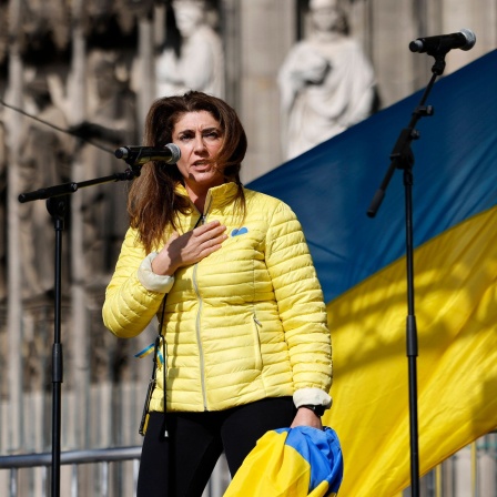 Linda Mai vom Blau-Gelben Kreuz steht bei einer Kundgebung am Mikrofon vor einer Ukraine-Flagge