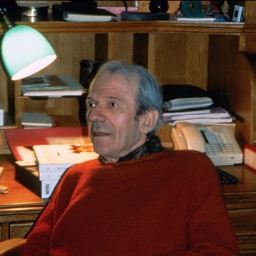 Der Philosoph Gilles Deleuze sitzt in einem rotem Pullover in Paris vor einem Schreibtisch. Im Hintergrund ist eine Lampe und ein Telefon.