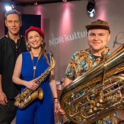 Eine Frau steht zwischen zwei Männern, alle lächeln. Ein Mann hält eine Tuba in der Hand und steht im Vordergrund, die Frau hält ein Saxofon in der Hand.