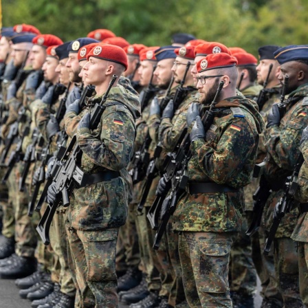 Eine Gruppe von Bundeswehrsoldaten in Tarnuniform und mit roten Baretten steht während einer Zeremonie mit Gewehren im Anschlag in einer Kaserne.