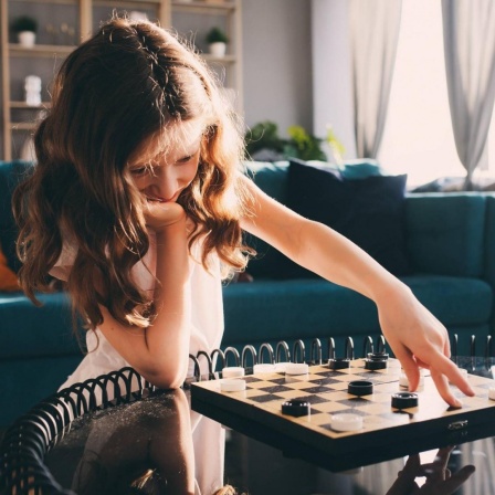 Ein Mädchen verschiebt mit der linken Hand einen Spielstein auf einem Brettspiel, ihr Kinn hat sie auf die rechte Hand gestützt.