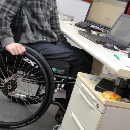 Ein Mann sitzt in einem Rollstuhl an seinem Arbeitsplatz am Schreibtisch. 