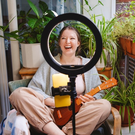 Blick durch ein Ringlicht mit daran montiertem Smartphone: Ein junge Frau sitzt lachend mit einer Ukulele auf einem Balkon